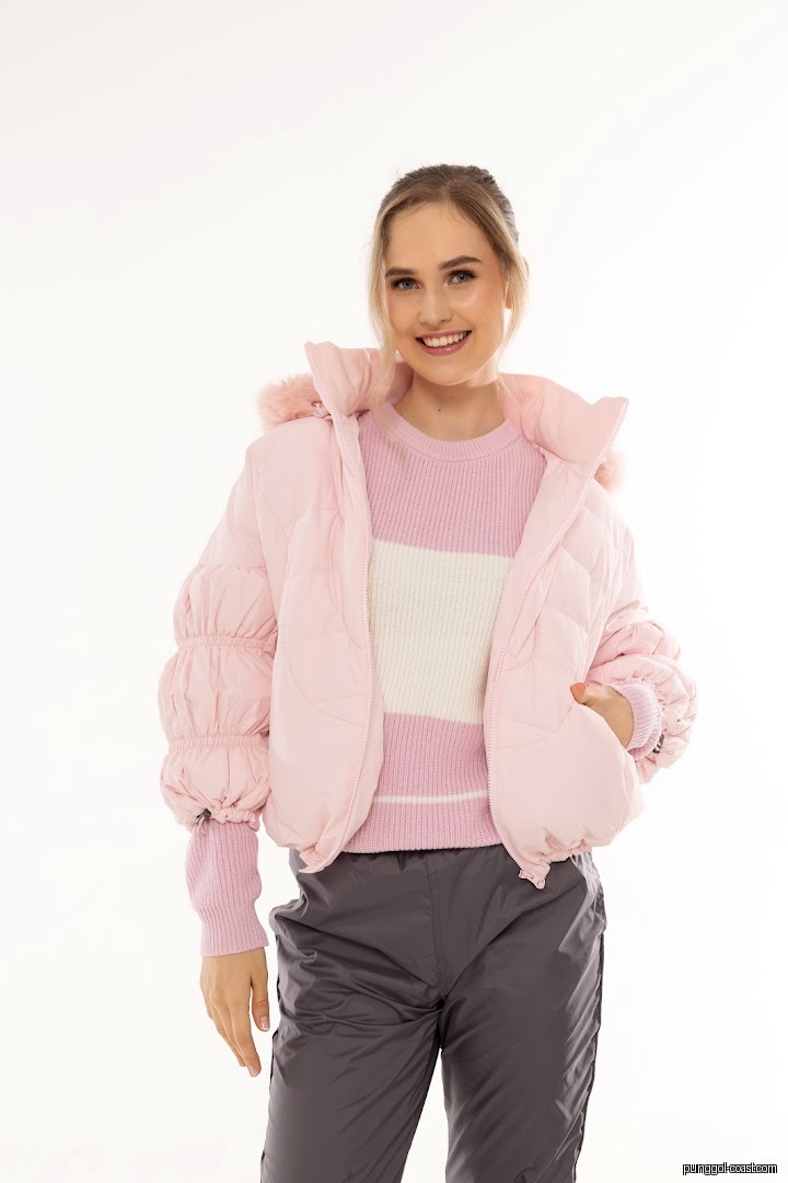 Woman models pink puffer jacket, pink sweater, and grey pants on white background.
