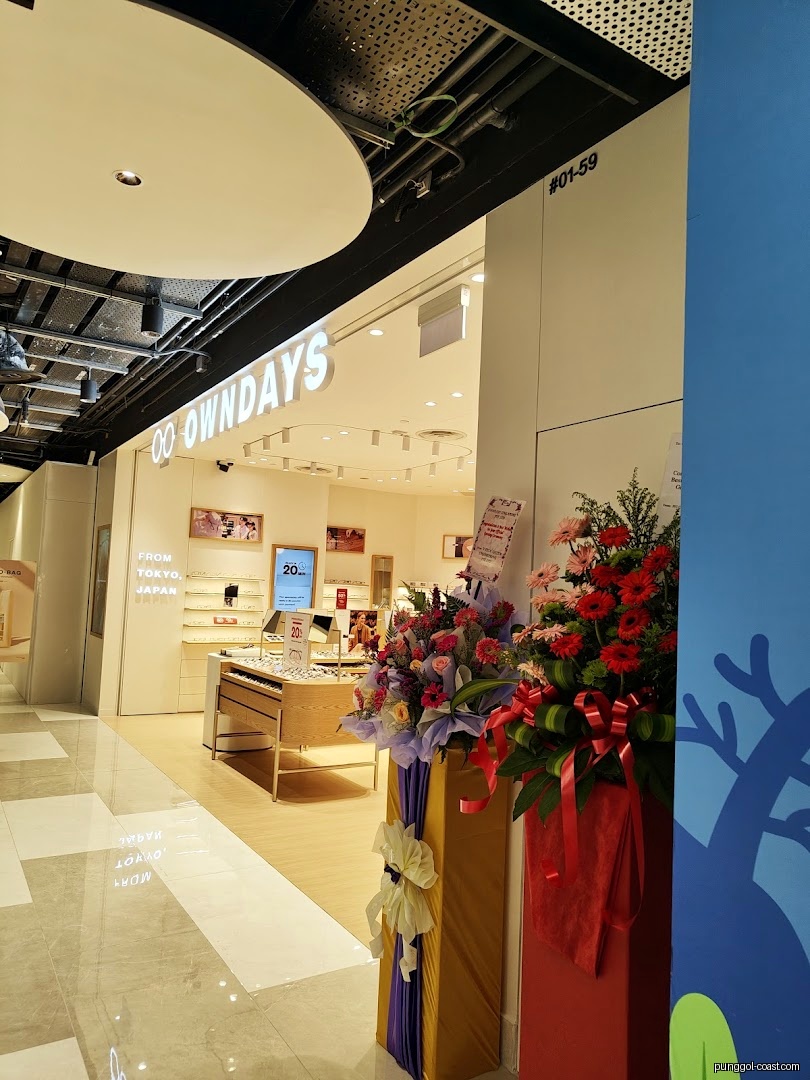 Entrance of OWNDAYS shop with flower arrangements in a mall.
