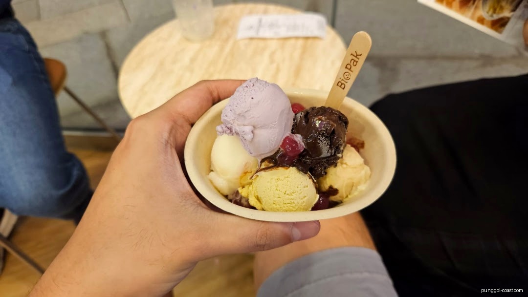 A hand holds a bowl of multi-scoop ice cream with a wooden spoon.