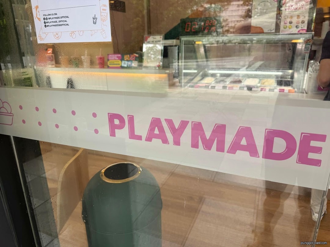 View through glass window into PlayMade shop, showing counter and ice cream display.