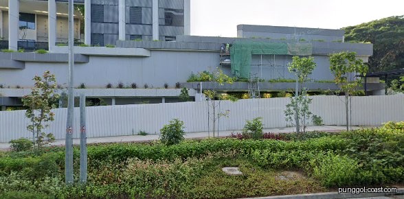 Exterior view of a building under construction, with a fence and lush greenery.