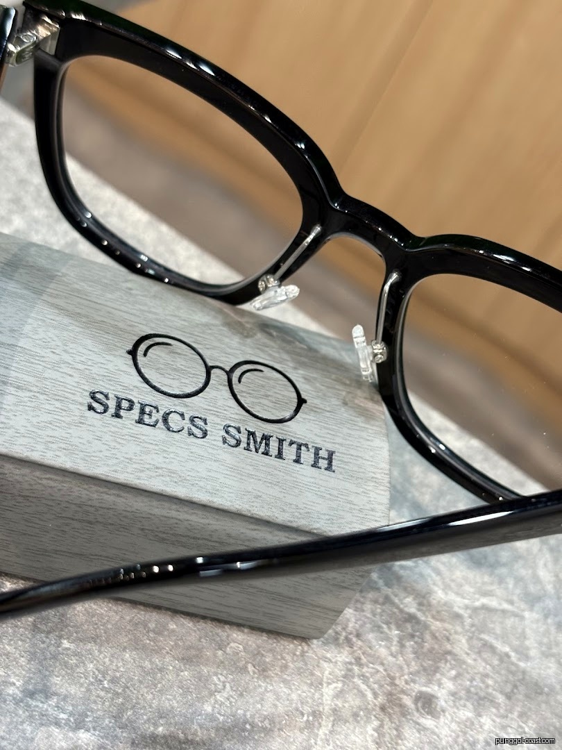 Black eyeglasses displayed on a wooden stand with 'Specs Smith' logo.