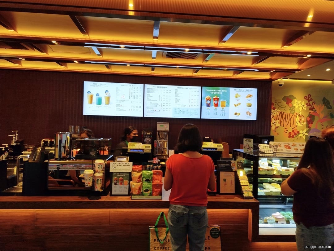 Customers ordering at a Starbucks counter with digital menus and a pastry display.