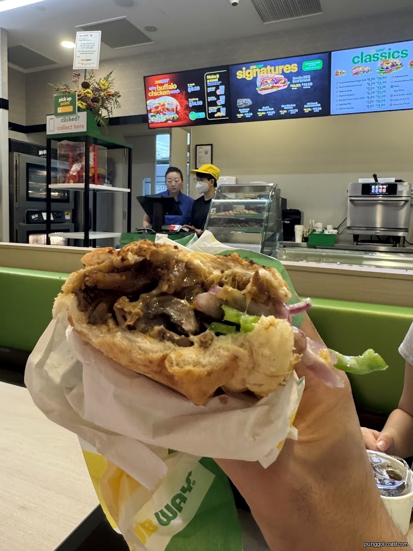 Person holding a Subway sandwich inside the shop with staff and menu boards.