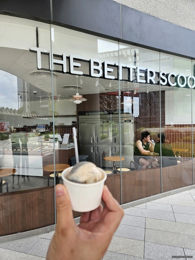 Hand holding ice cream cup in front of 'The Better Scoop' storefront.