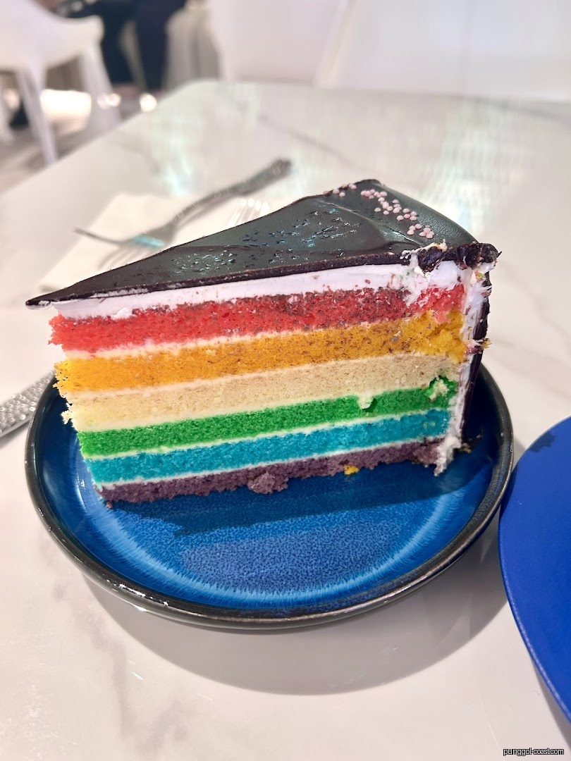 A colorful slice of rainbow cake with chocolate frosting on a blue plate.