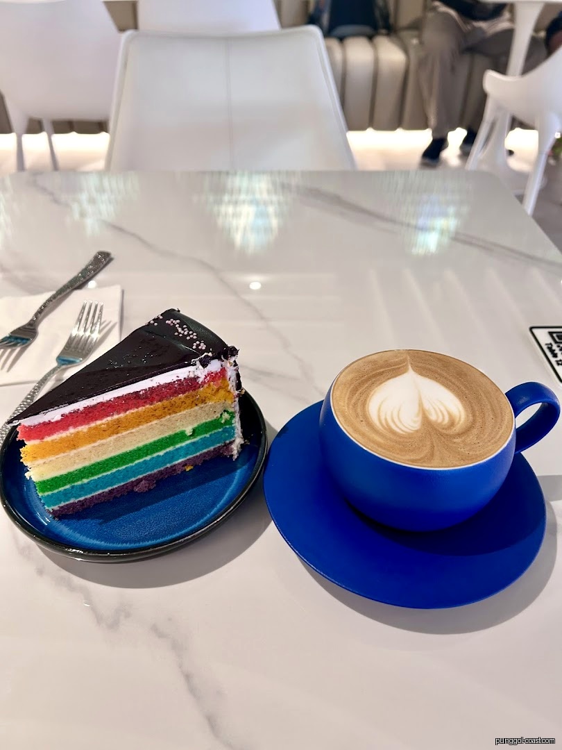 A slice of rainbow cake and a blue cup of coffee on a white table.