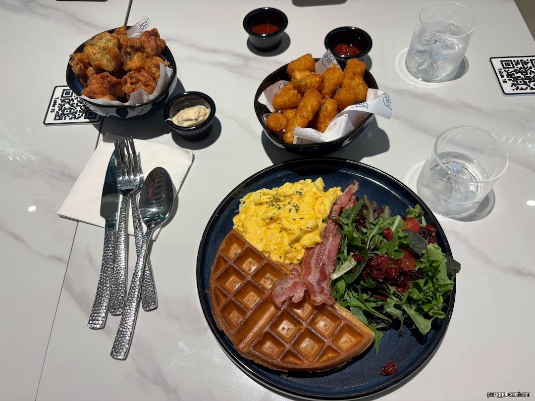 A table set with a meal of waffles, eggs, bacon, salad, and fried snacks.