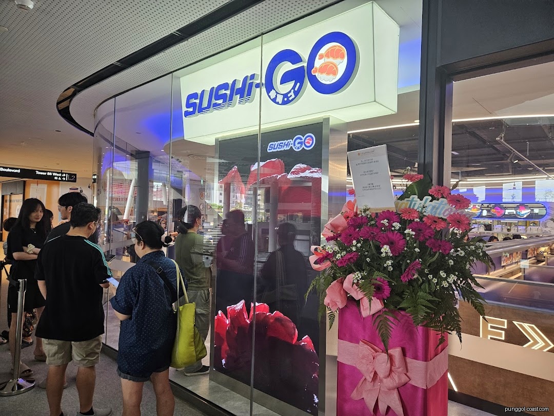 Sushi-GO shop front with people waiting and a large floral display.